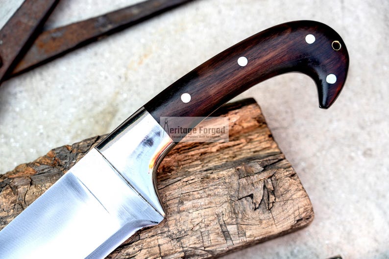 May include: A close-up of a large, silver knife with a dark wood handle. The handle has white dots and a gold ring. The knife is resting on a piece of wood.