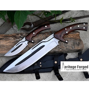 May include: Two knives with wooden handles and a black leather sheath. The larger knife has a curved blade and a decorative design on the blade. The smaller knife has a straight blade. The knives are on a wooden surface. The text "Heritage Forged" is visible on the sheath.  Forged with Passion, Built for Legends.