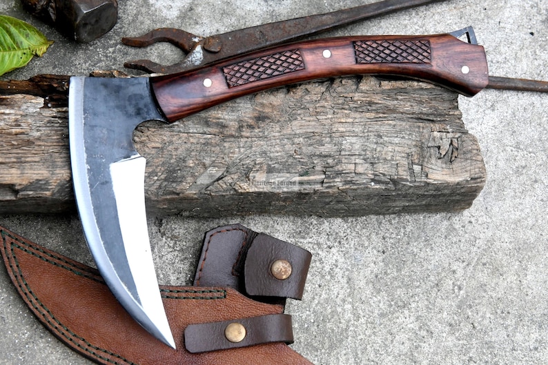 May include: A handcrafted axe with a dark wooden handle and a curved, silver blade. The handle features decorative diamond-shaped carvings. The axe rests on a weathered wooden block, with a brown leather sheath and other tools in the background.