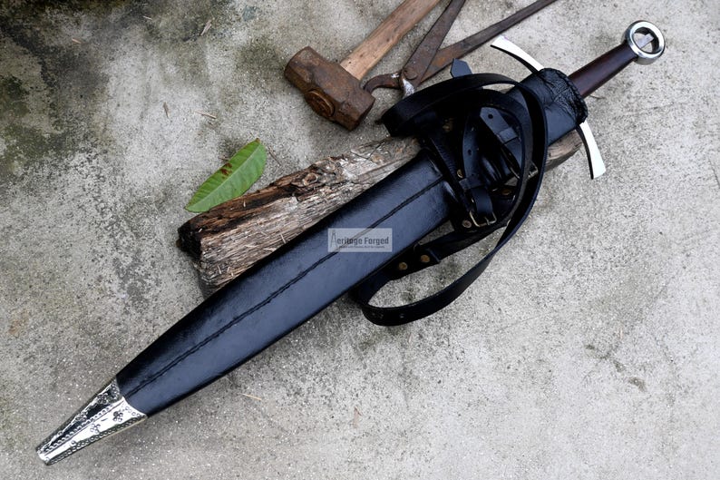 Op de afbeelding: Een zwarte leren schede met een zilverkleurig zwaard erin. Het zwaard heeft een zilveren knop en een zwarte leren greep. De schede is bevestigd aan een bruine leren riem.