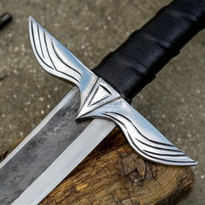 May include: Close-up of a silver and black sword with a leather-wrapped handle. The blade is silver and the guard has a wing-like design. The sword rests on a piece of wood.