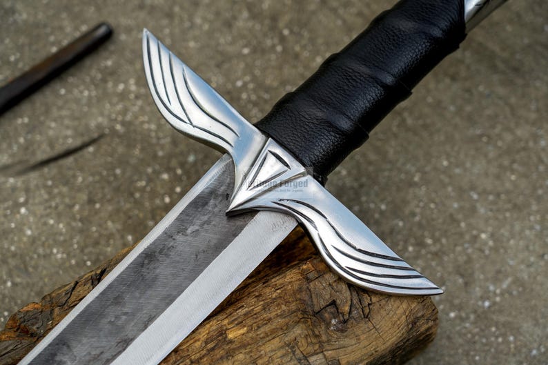 May include: Close-up of a silver and black sword with a leather-wrapped handle. The blade is silver and the guard has a wing-like design. The sword rests on a piece of wood.