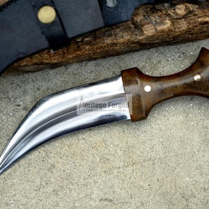 May include: A curved, silver-coloured dagger with a brown wooden handle. The handle has a white dot on the top. The dagger is lying on a grey, textured surface. The text "Heritage Forged" is engraved on the blade.