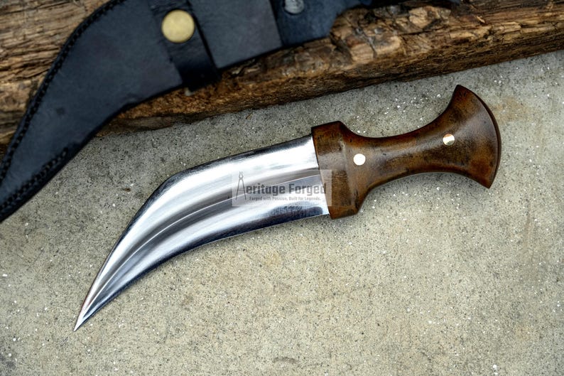 May include: A curved, silver-coloured dagger with a brown wooden handle. The handle has a white dot on the top. The dagger is lying on a grey, textured surface. The text "Heritage Forged" is engraved on the blade.