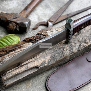 Puede incluir: Una hoja larga de plata con un mango de madera marr&oacute;n y una funda de cuero. La hoja es afilada y puntiaguda, y el mango est&aacute; decorado con un patr&oacute;n. El cuchillo est&aacute; apoyado en un trozo de madera.
