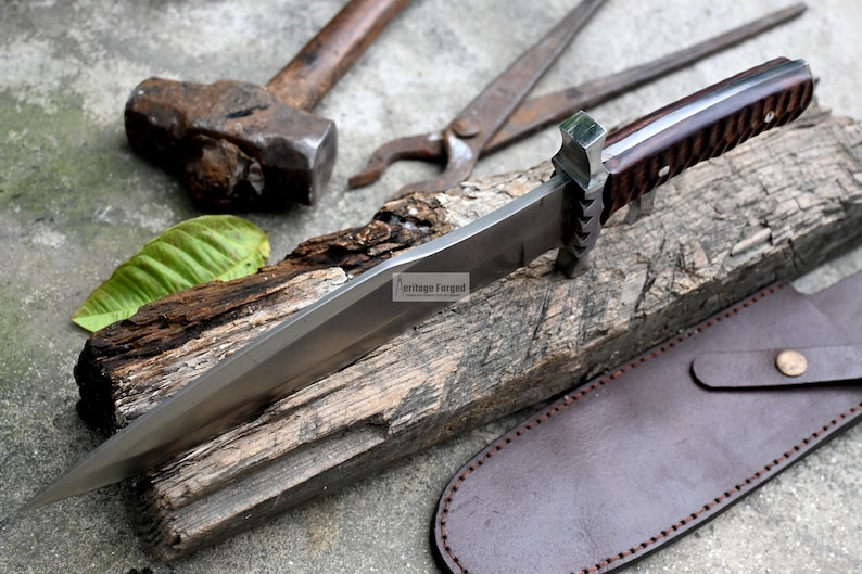 Puede incluir: Una hoja larga de plata con un mango de madera marr&oacute;n y una funda de cuero. La hoja es afilada y puntiaguda, y el mango est&aacute; decorado con un patr&oacute;n. El cuchillo est&aacute; apoyado en un trozo de madera.