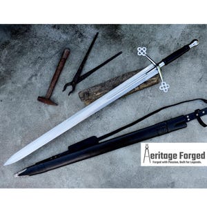 Op de afbeelding: Een lang, zilveren zwaard met een zwarte handgreep en pareerstang, rustend op een houten plank. Een zwarte leren schede en andere smidgereedschappen zijn ook aanwezig. De tekst "Aeritage Forged" is zichtbaar.