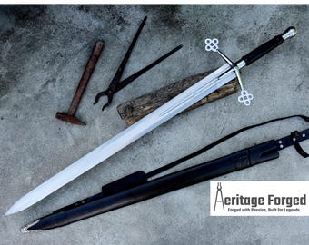 Puede incluir: Una espada larga y plateada con empuñadura y guarda negras, apoyada sobre una tabla de madera. También hay una funda de cuero negro y otras herramientas de herrero. El texto "Aeritage Forged" es visible.