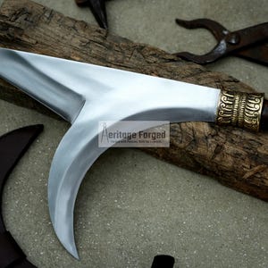 May include: A curved, silver-colored blade with a wooden handle and a brass-colored decorative piece. The blade is resting on a piece of wood.