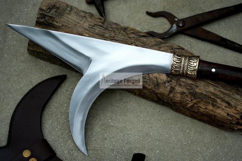May include: A curved, silver-colored blade with a wooden handle and a brass-colored decorative piece. The blade is resting on a piece of wood.