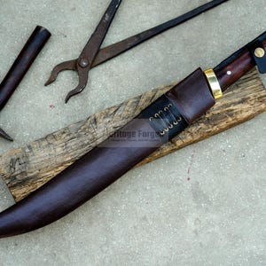 May include: A brown leather scabbard with a curved blade and a wooden handle. The scabbard has a brass tip and a brass button. The blade is sharp and has a slight curve. The handle is made of dark wood and has a brass pommel. The scabbard is lying on a wooden surface. The text "Heritage Forge" is visible on the scabbard.