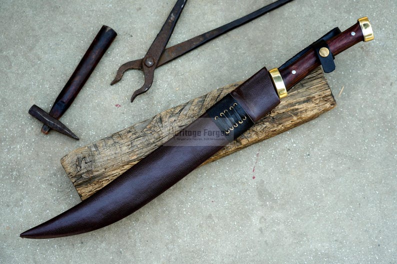 May include: A brown leather scabbard with a curved blade and a wooden handle. The scabbard has a brass tip and a brass button. The blade is sharp and has a slight curve. The handle is made of dark wood and has a brass pommel. The scabbard is lying on a wooden surface. The text "Heritage Forge" is visible on the scabbard.
