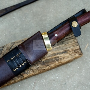 May include: A brown leather sheath with a black leather strap and brass accents. The sheath is holding a knife with a dark wood handle and a brass pommel.