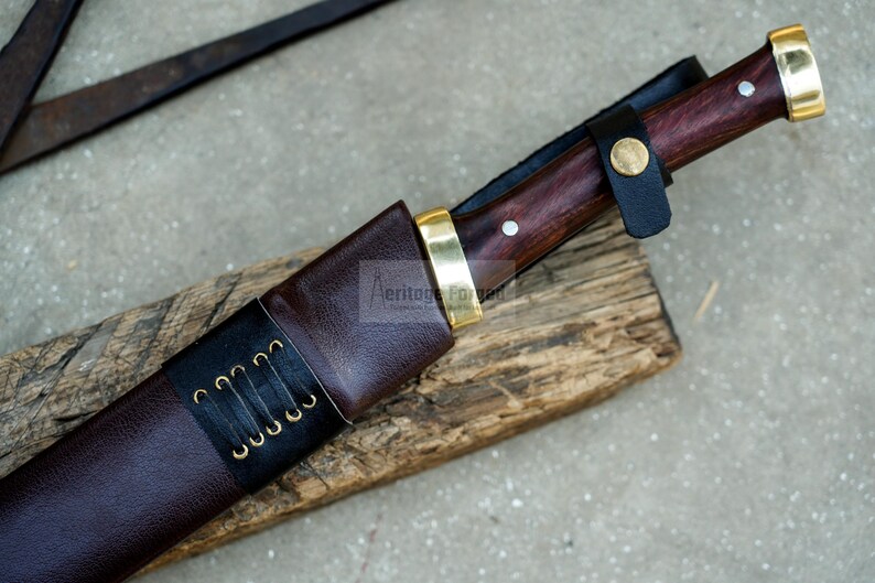 May include: A brown leather sheath with a black leather strap and brass accents. The sheath is holding a knife with a dark wood handle and a brass pommel.