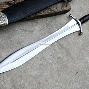 May include: A silver-coloured sword with a leather-wrapped handle and a silver pommel. The sword is lying on a grey surface with a wooden scabbard in the background.