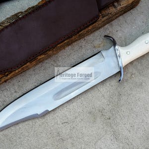 Op de afbeelding: Een groot, zilverkleurig Bowie-mes met een witte handgreep en een leren schede. Het mes is scherp en heeft een puntige punt. De schede is gemaakt van bruin leer en heeft een klep die het mes vastzet.