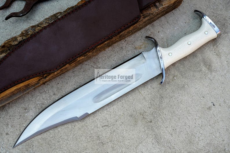 Op de afbeelding: Een groot, zilverkleurig Bowie-mes met een witte handgreep en een leren schede. Het mes is scherp en heeft een puntige punt. De schede is gemaakt van bruin leer en heeft een klep die het mes vastzet.