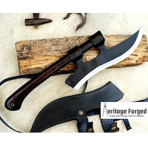 Op de afbeelding: Een zwarte en zilveren bijl met een donkerhouten handvat, rustend op een stuk hout. De bijl heeft een gebogen blad met een witte rand. Een zwarte leren schede bevindt zich eronder. De tekst "Aeritage Forged" is zichtbaar.