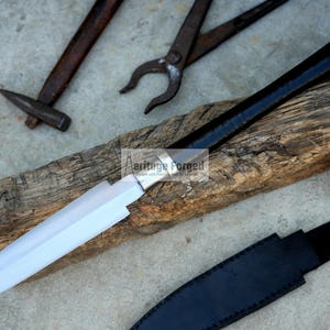 May include: A handcrafted spear with a polished blade and a black handle with a silver end cap rests on a piece of wood. A black leather sheath is visible, along with blacksmith tools in the background.