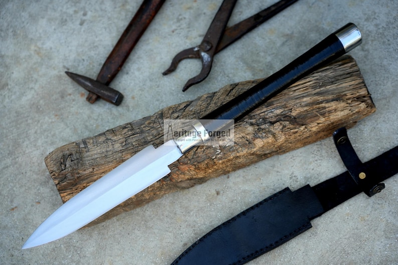 May include: A handcrafted spear with a polished blade and a black handle with a silver end cap rests on a piece of wood. A black leather sheath is visible, along with blacksmith tools in the background.