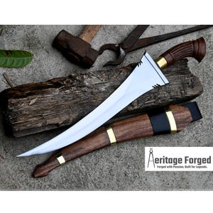 May include: A curved, silver blade with a wooden handle and gold accents. The blade is sharp and pointed, and the handle is decorated with a dark brown wood. The blade is resting on a wooden surface. The text "Heritage Forged" is visible in the lower right corner of the image. "Forged with Passion, Built for Legends."