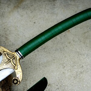 May include: A close-up of a sword with a green wrapped handle and a gold-colored hilt. The hilt features an ornate design with floral accents. The blade is silver with a black line down the center.