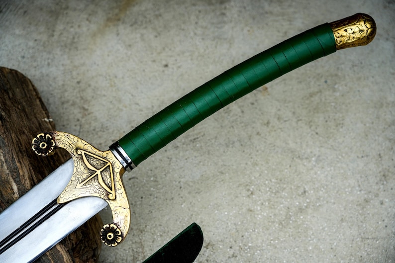 May include: A close-up of a sword with a green wrapped handle and a gold-colored hilt. The hilt features an ornate design with floral accents. The blade is silver with a black line down the center.