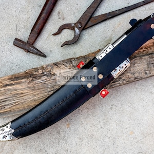 May include: A black leather-sheathed knife with a silver blade and decorative accents. The knife is resting on a piece of wood with the text "Heritage Forged" visible.
