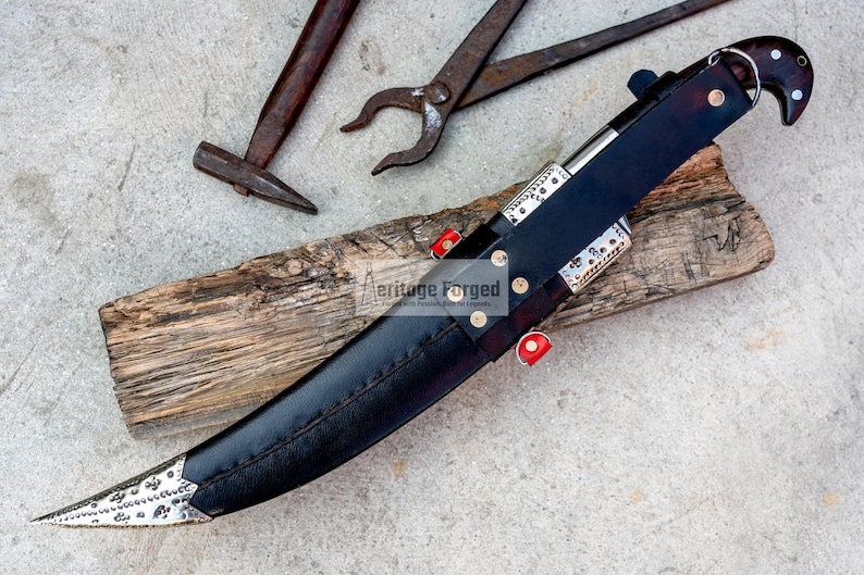 May include: A black leather-sheathed knife with a silver blade and decorative accents. The knife is resting on a piece of wood with the text "Heritage Forged" visible.