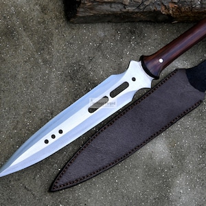 May include: A silver-colored spear with a brown wooden handle and a leather sheath. The spear has a long, pointed blade with a series of holes along the edge.