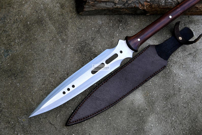 May include: A silver-colored spear with a brown wooden handle and a leather sheath. The spear has a long, pointed blade with a series of holes along the edge.