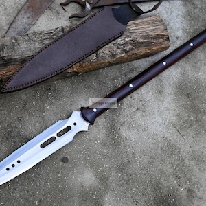 May include: A long, silver-colored spear with a dark brown wooden handle. The spear has a sharp point and a decorative design on the blade. The spear is resting on a gray surface.