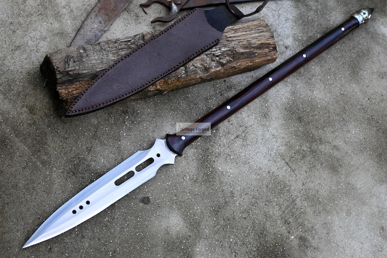 May include: A long, silver-colored spear with a dark brown wooden handle. The spear has a sharp point and a decorative design on the blade. The spear is resting on a gray surface.