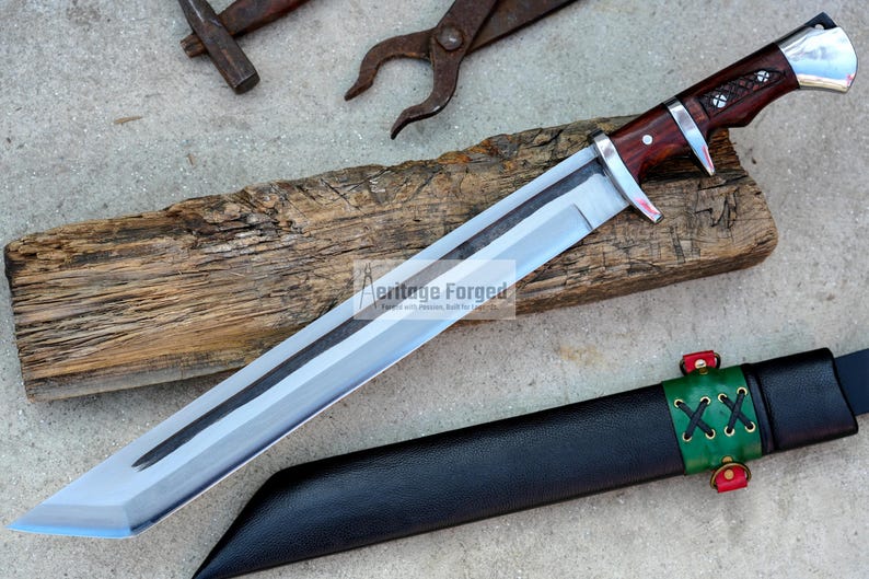 May include: A silver and brown knife with a wooden handle and a black leather sheath. The knife has a long, sharp blade and a decorative design on the handle. The sheath has a green strap with a double X design and red accents.