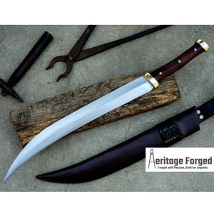 May include: A curved, silver-colored sword with a brown wooden handle and a leather sheath. The sword is resting on a wooden surface. The text "Heritage Forged" is visible in the lower right corner of the image. "Forged with Passion, Built for Legends."