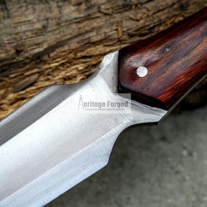 K&ouml;nnte beinhalten: Nahaufnahme einer silbernen Messerklinge mit einem Holzgriff. Der Griff ist dunkelbraun mit einem r&ouml;tlichen Farbton und hat zwei silberne Nieten. Die Klinge ist scharf und hat eine leichte Kr&uuml;mmung. Das Messer liegt auf einem St&uuml;ck Holz. Der Text "Heritage Forged" ist auf der Klinge sichtbar.