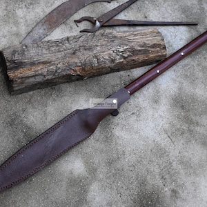May include: A long, brown leather-wrapped knife with a silver tip and a wooden handle. The knife is lying on a concrete surface with a piece of wood and a metal tool in the background.