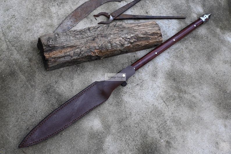 May include: A long, brown leather-wrapped knife with a silver tip and a wooden handle. The knife is lying on a concrete surface with a piece of wood and a metal tool in the background.