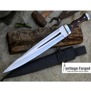 May include: A long, silver-colored dagger with a dark wood handle and a black leather sheath. The dagger has a single-edged blade and a pointed tip. The sheath has a decorative strap with stitching. The text "Heritage Forged" is visible in the lower right corner of the image. "Forged with Passion, Built for Legends."