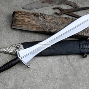 May include: A silver sword with a black leather scabbard and a silver pommel. The sword has a decorative pattern on the pommel and scabbard. The sword is on a grey surface with a piece of wood and a pair of pliers.