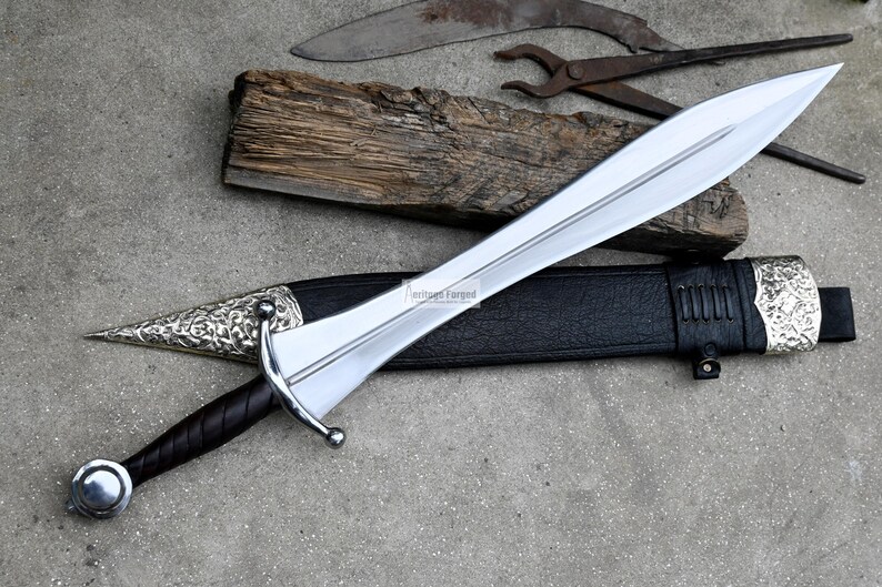 May include: A silver sword with a black leather scabbard and a silver pommel. The sword has a decorative pattern on the pommel and scabbard. The sword is on a grey surface with a piece of wood and a pair of pliers.