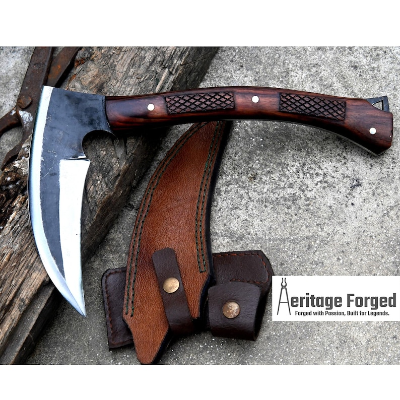 May include: Axe with a curved blade and a dark wooden handle. The handle features a diamond pattern and is secured with small rivets. A brown leather sheath with a belt loop protects the blade. The text "Aeritage Forged" is visible.