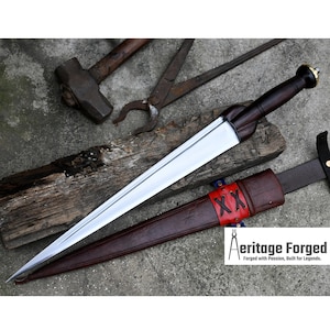 May include: A silver dagger with a dark wood handle and a leather scabbard with red stitching. The scabbard has the text "XX" on it. The dagger is resting on a wooden surface.