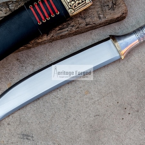 May include: A silver and gold curved dagger with a decorative handle. The blade is sharp and the handle has intricate details. The dagger is on a gray surface. The text "Heritage Forged" is visible on the blade.