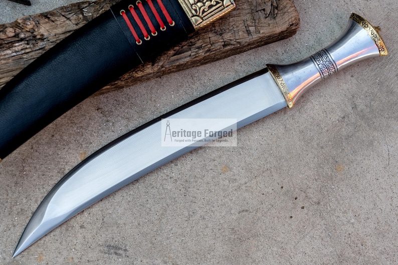 May include: A silver and gold curved dagger with a decorative handle. The blade is sharp and the handle has intricate details. The dagger is on a gray surface. The text "Heritage Forged" is visible on the blade.