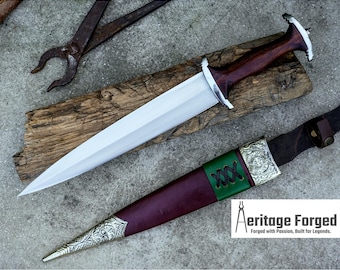 May include: A handcrafted dagger with a polished steel blade and a dark wood handle, resting on a wooden plank. The dagger is accompanied by a burgundy and green leather sheath with silver-colored metal accents. The text "Aeritage Forged" is visible.