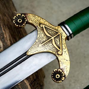 May include: Close-up of a decorative sword with a silver blade and a gold-colored hilt. The hilt features an intricate floral design and a green handle. The sword is resting on a wooden surface.