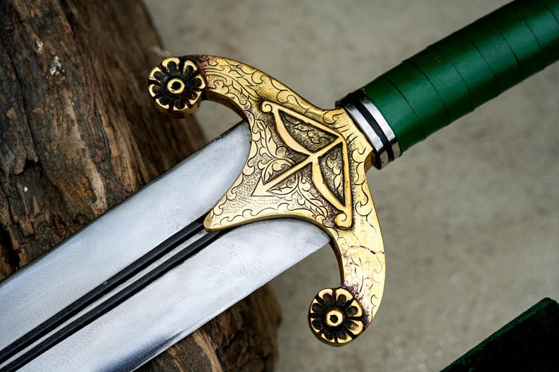 May include: Close-up of a decorative sword with a silver blade and a gold-colored hilt. The hilt features an intricate floral design and a green handle. The sword is resting on a wooden surface.