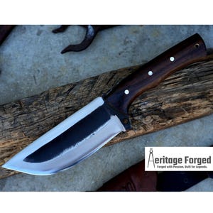 May include: A large, black-handled knife with a silver blade. The knife is resting on a wooden surface. The text "Heritage Forged" is visible in the lower right corner of the image. "Forged with Passion, Built for Legends."