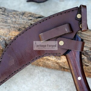 Op de afbeelding: Een bruine leren schede met een bruine houten handvat en een metalen mes. De schede heeft een riem en een metalen klinknagel. De tekst "Heritage Forged" is op de schede gedrukt.